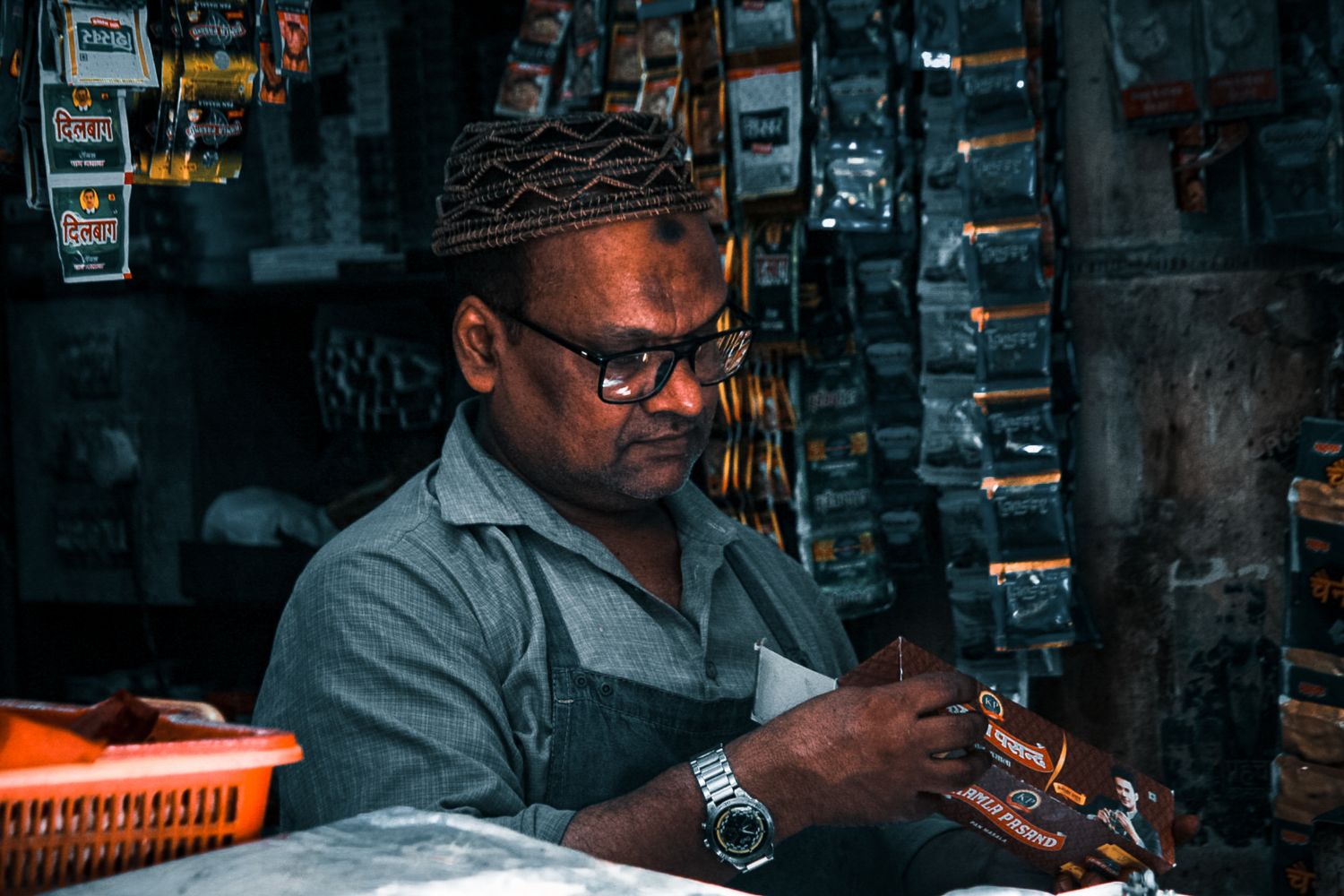 A mature South Asian shopkeeper organizing products in his store, wearing glasses and a patterned cap.