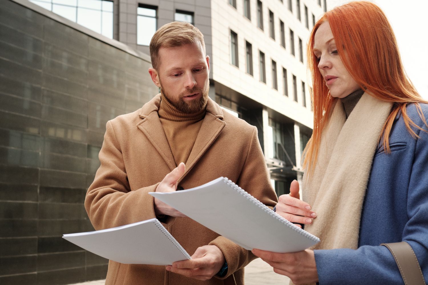 Two business professionals in coats discuss documents in an urban setting.